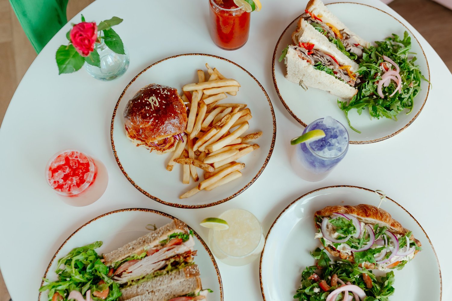 Four brunch plates on a white table: burger and fries, club sandwiches and salads